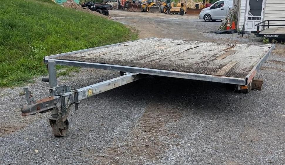 Flatbed trailer with wooden deck parked on gravel near a grassy roadside.