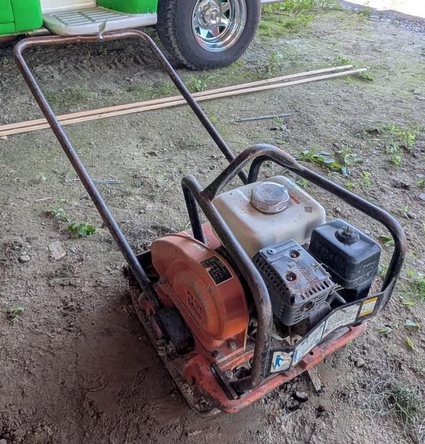 Small red plate compactor on dirt, with a gas engine and long handle outdoors near a vehicle.