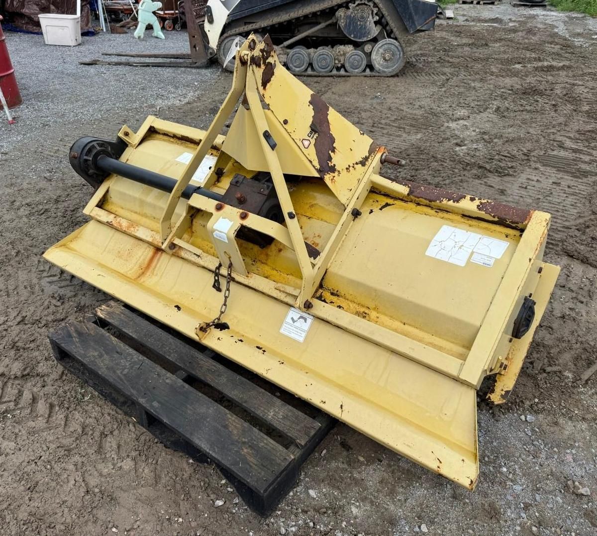 Yellow rotary mower attachment on gravel, with black side panel and hydraulic arm in a farmyard.