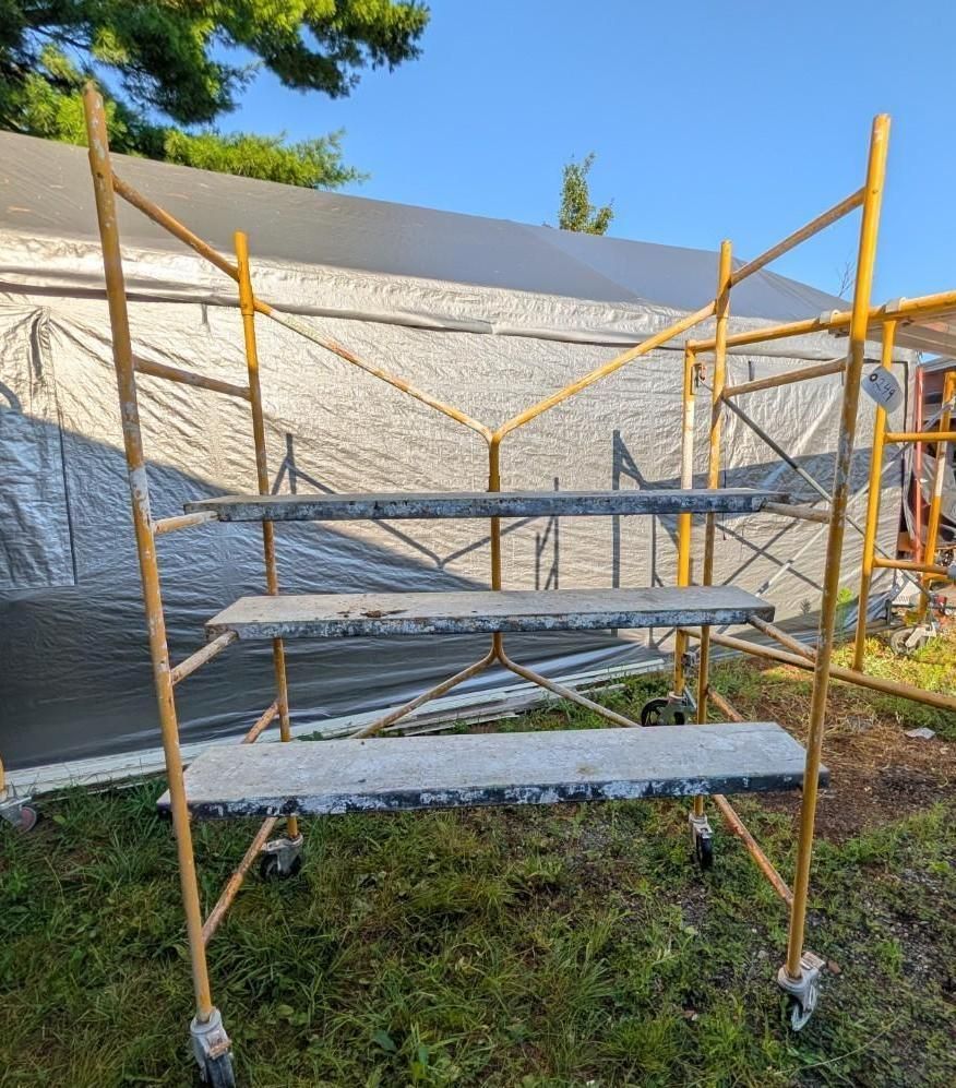 Yellow scaffolding on wheels around a small raised work platform outdoors against a tarp-covered wall