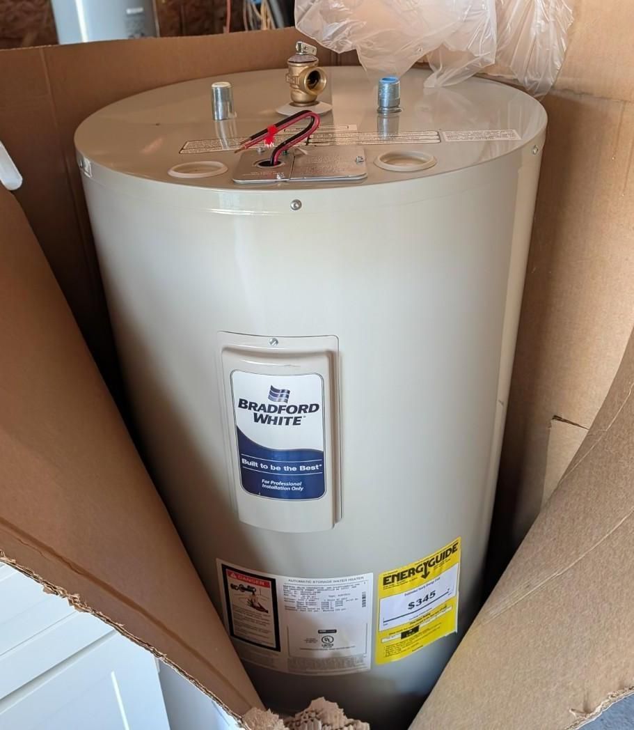 White water heater in a cardboard box, with pipes and labels visible on top and front.
