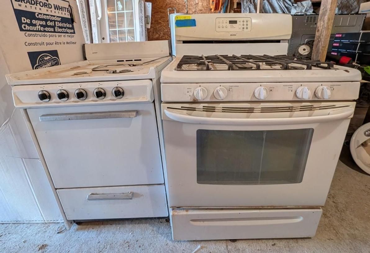 Two white kitchen appliances: a stove and a dishwasher, shown side by side in a garage.