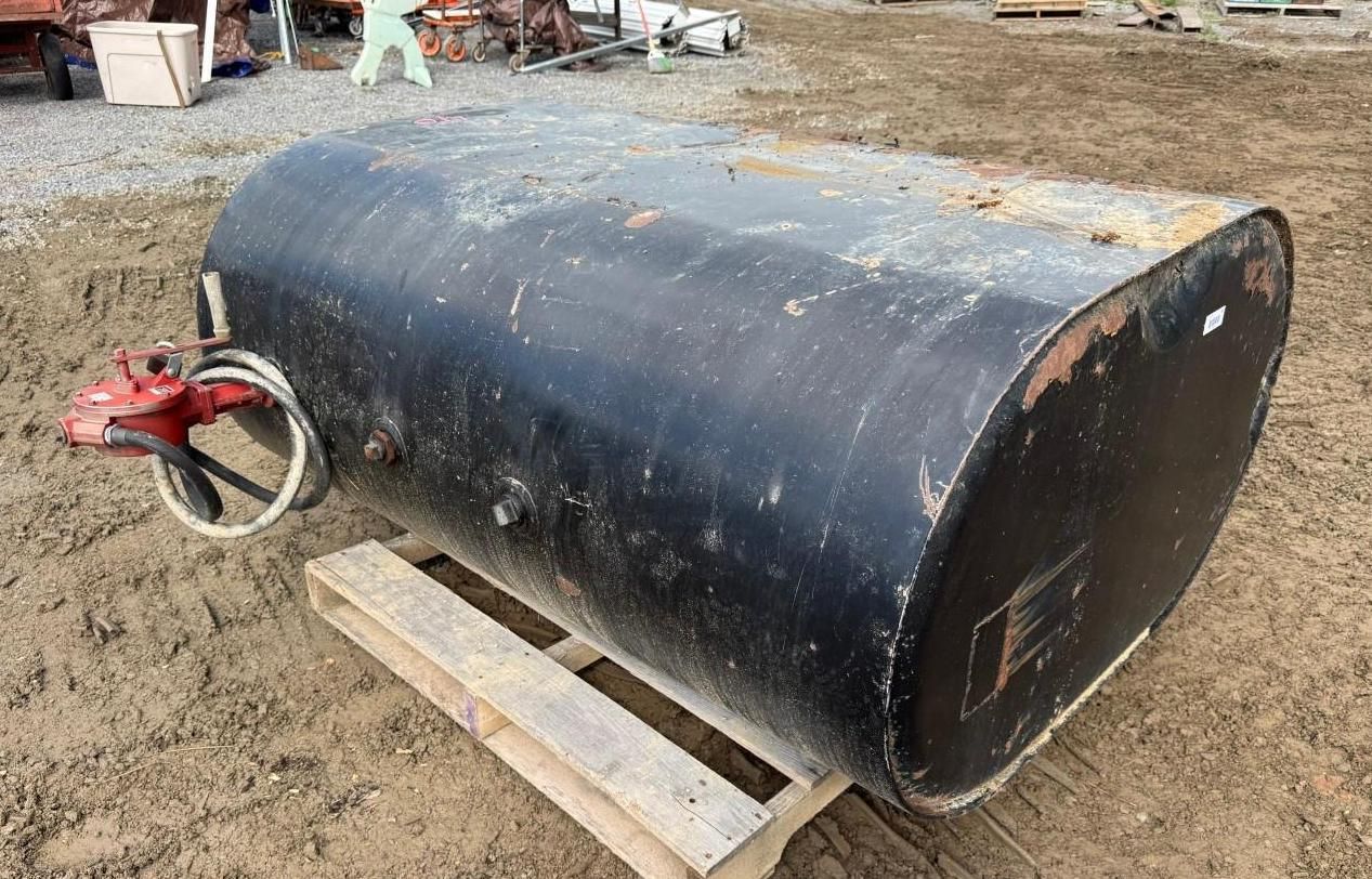 Large black cylindrical tank on wooden blocks in a muddy yard, with a red valve on one end