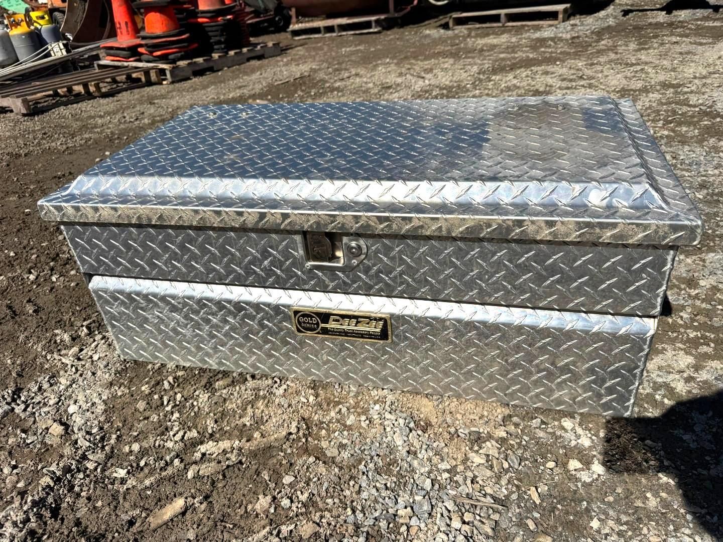 Silver diamond-plate metal toolbox with lid, sitting on gravel outdoors.