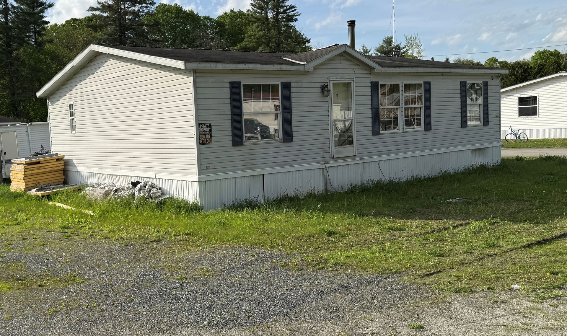 White single-story mobile home with windows and porch on a grassy gravel lot under a sunny sky