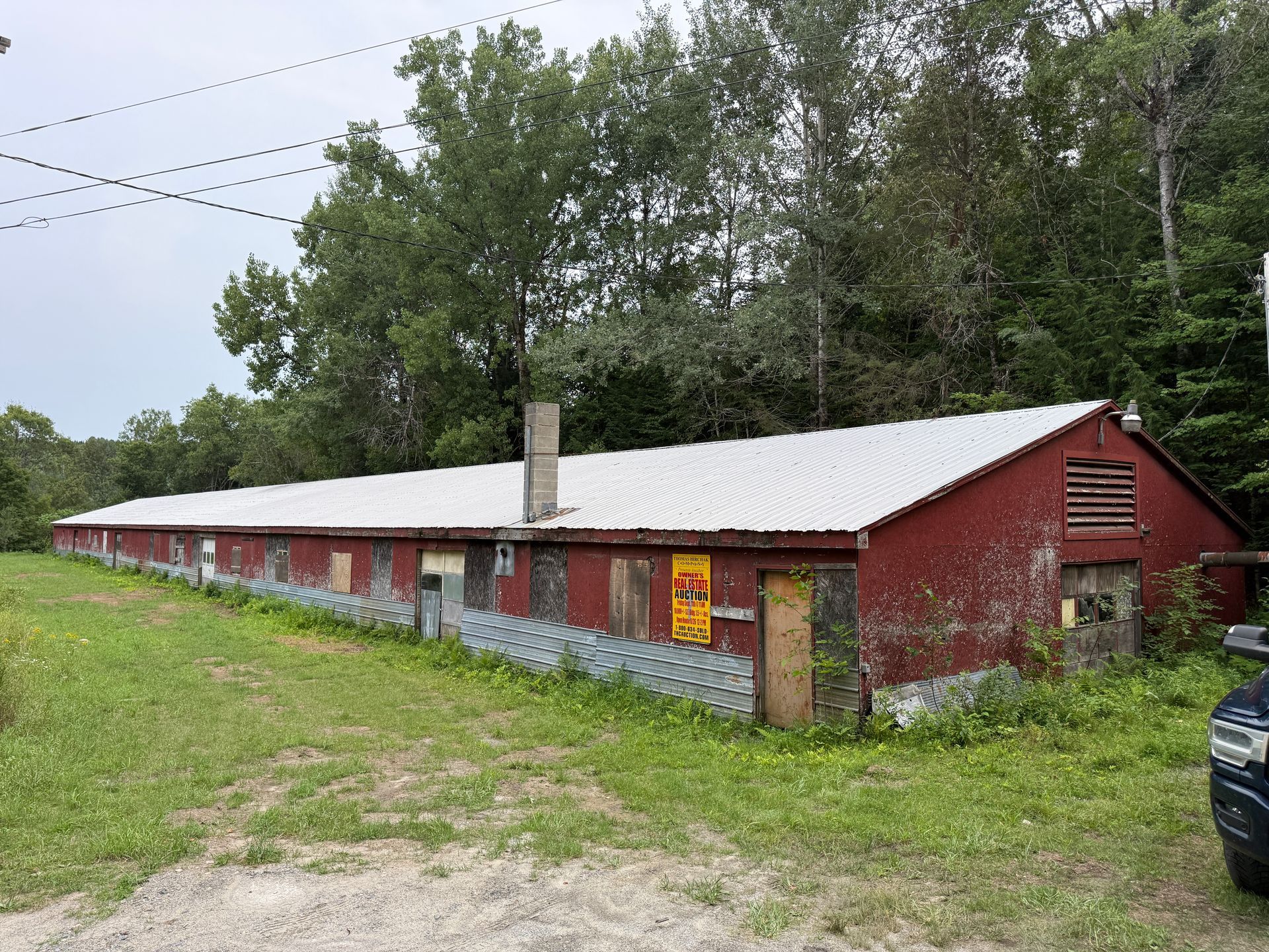 Long red wooden building with a white roof in a grassy rural area, trees behind it.