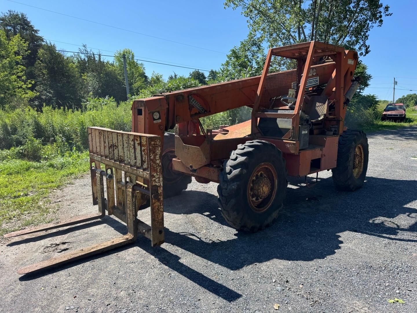 orange forklift with raised forks parked on a gravel road in a sunny wooded area