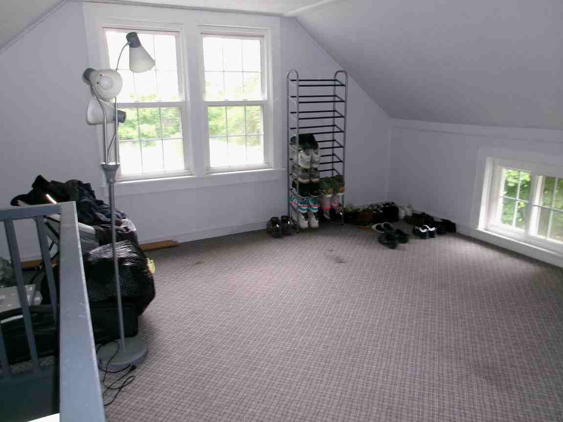 Empty carpeted attic room with slanted ceiling, window seats, shoe rack, and a standing fan near a window