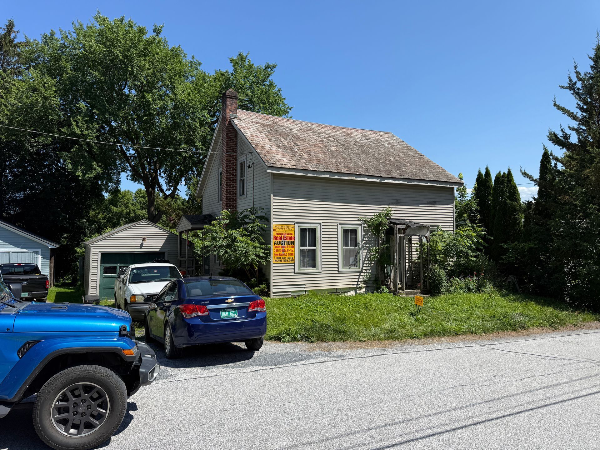 Small white house with a front porch beside parked cars on a sunny residential street