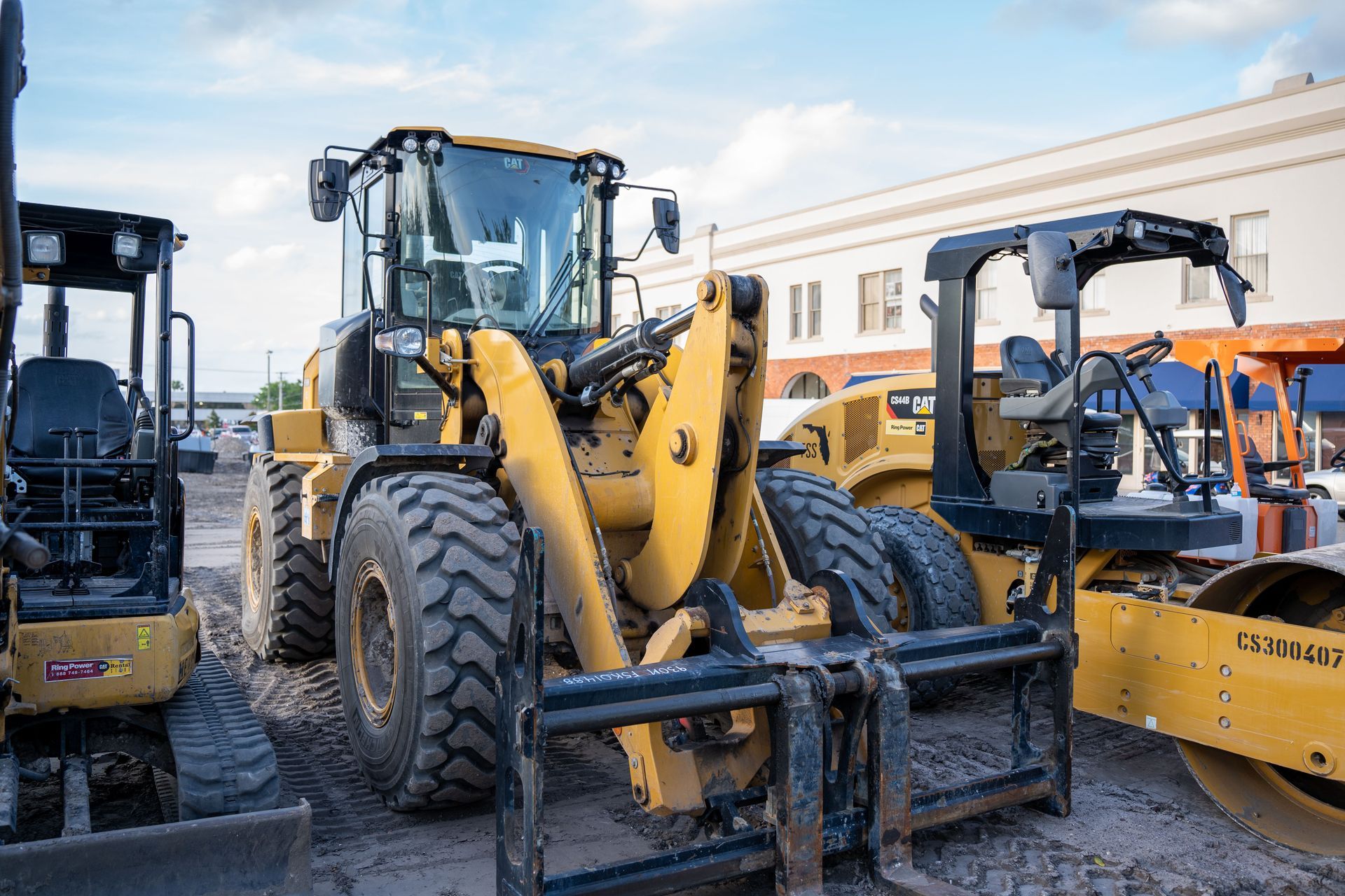 Yellow construction vehicles parked on a gravel lot beside a building under a blue sky