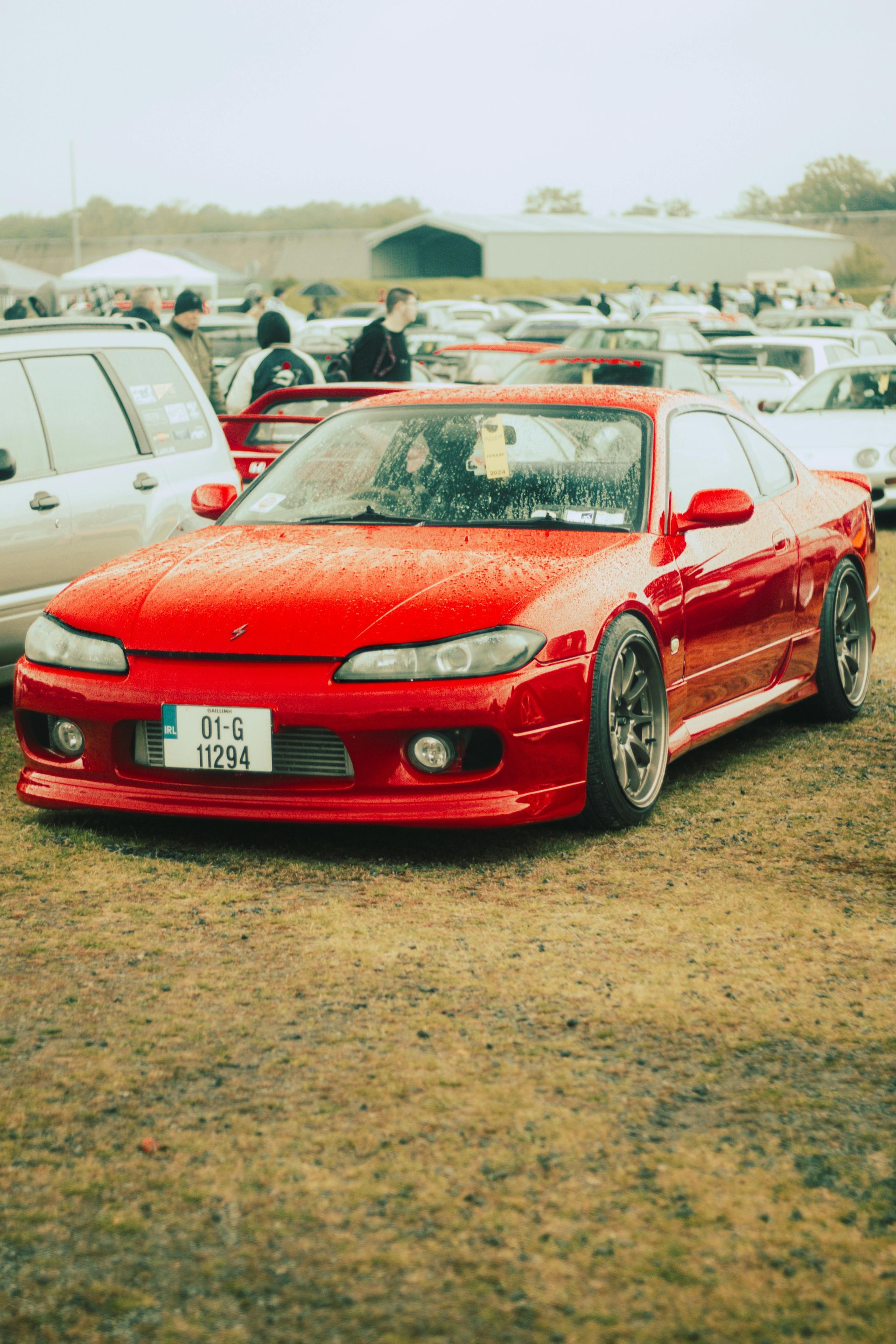 Red sports car parked on a dirt lot at an outdoor car meet, with other vehicles in the background.