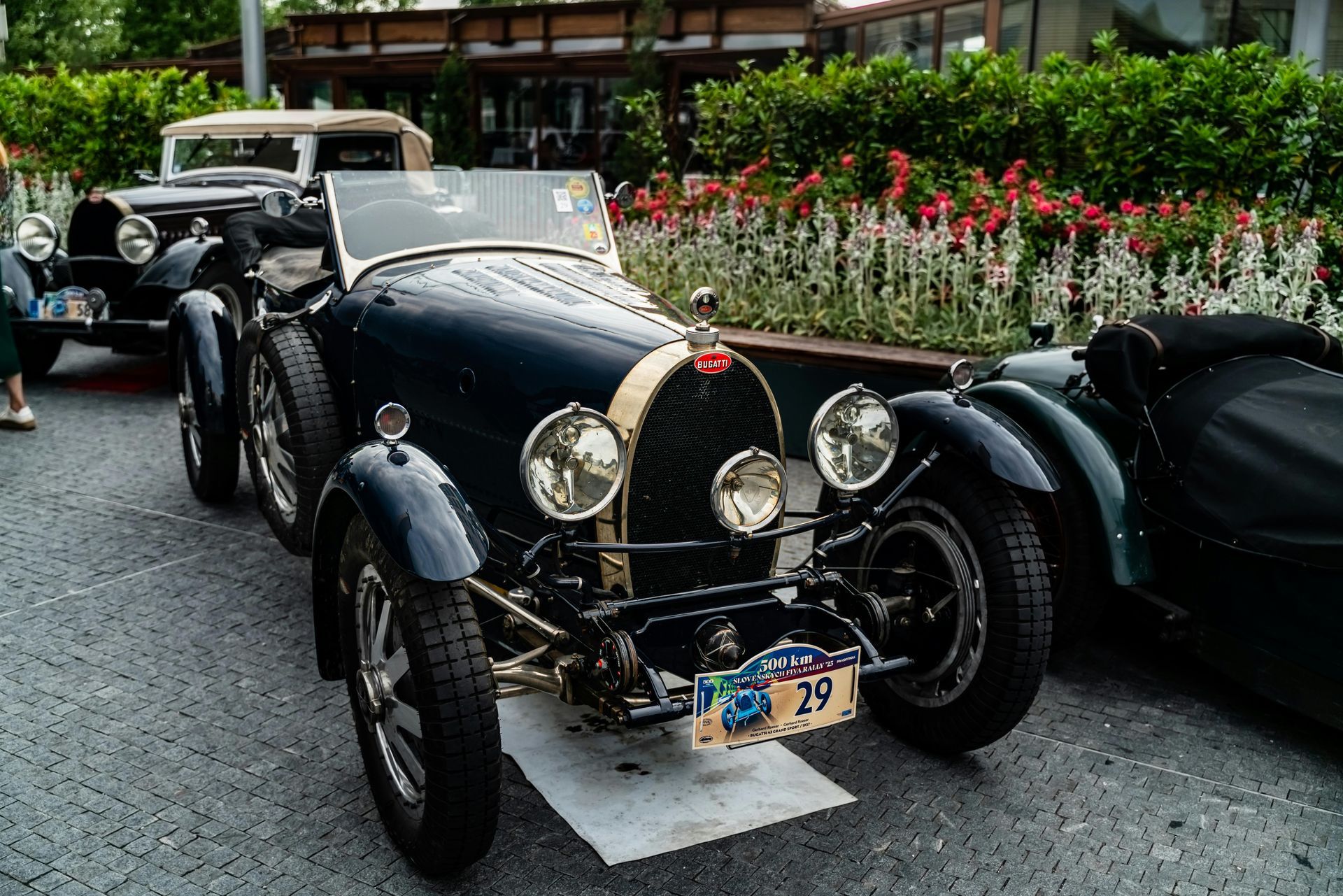 Vintage black Bugatti parked outdoors beside flowers, with another classic car behind it.
