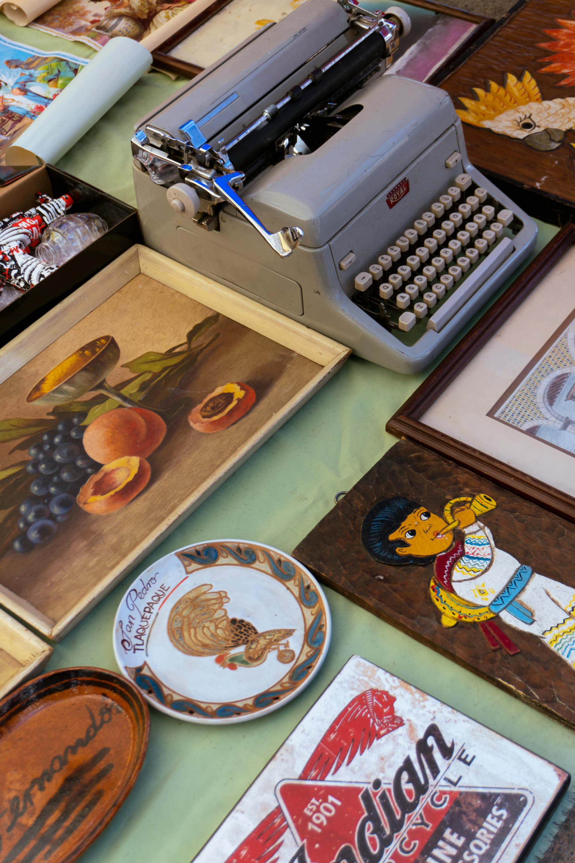 Assorted vintage framed art and a typewriter arranged on a table, with colorful plates and decor