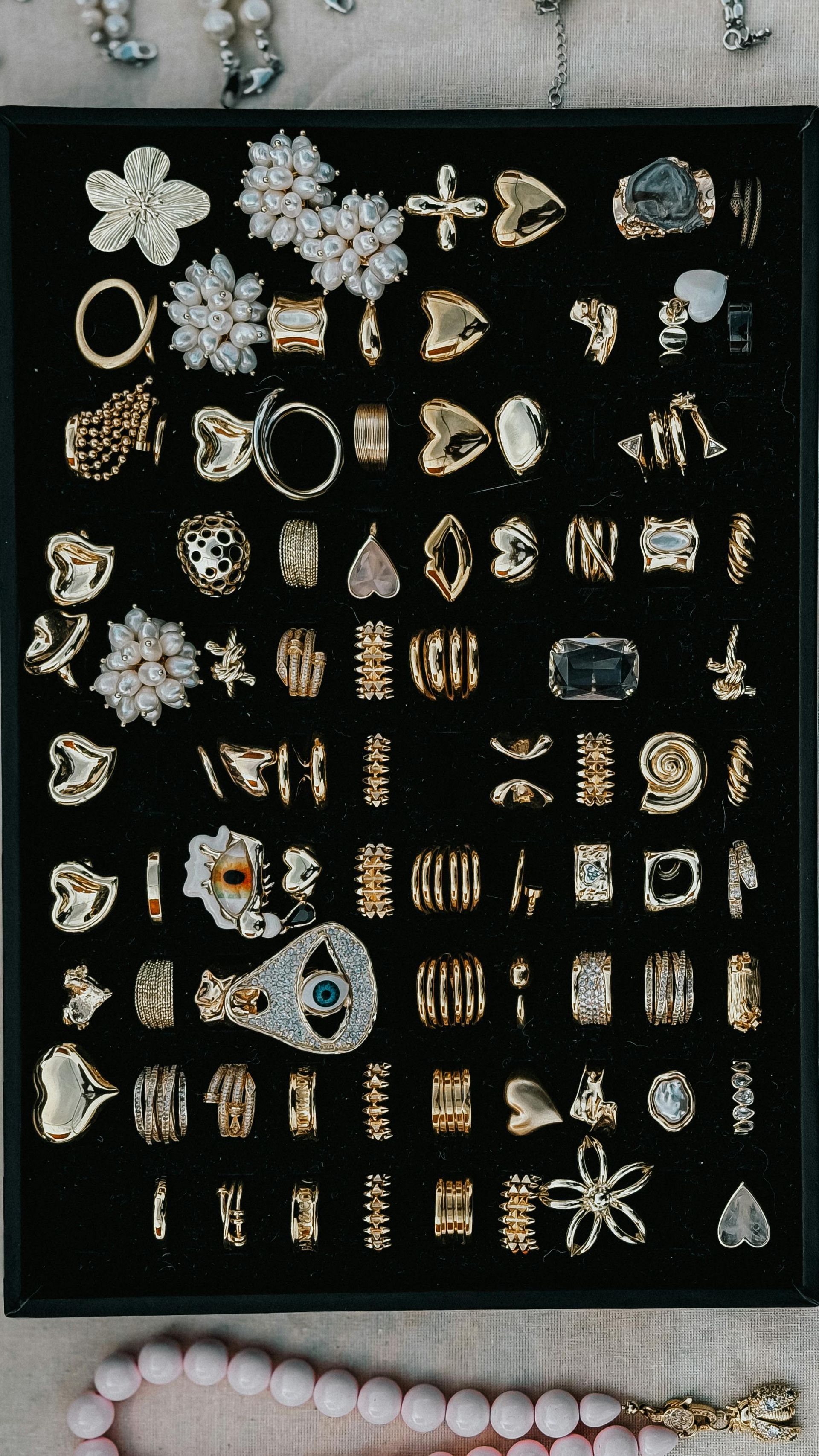 Assorted gold and silver rings arranged on black display boards, with a pink beaded necklace partly visible below.