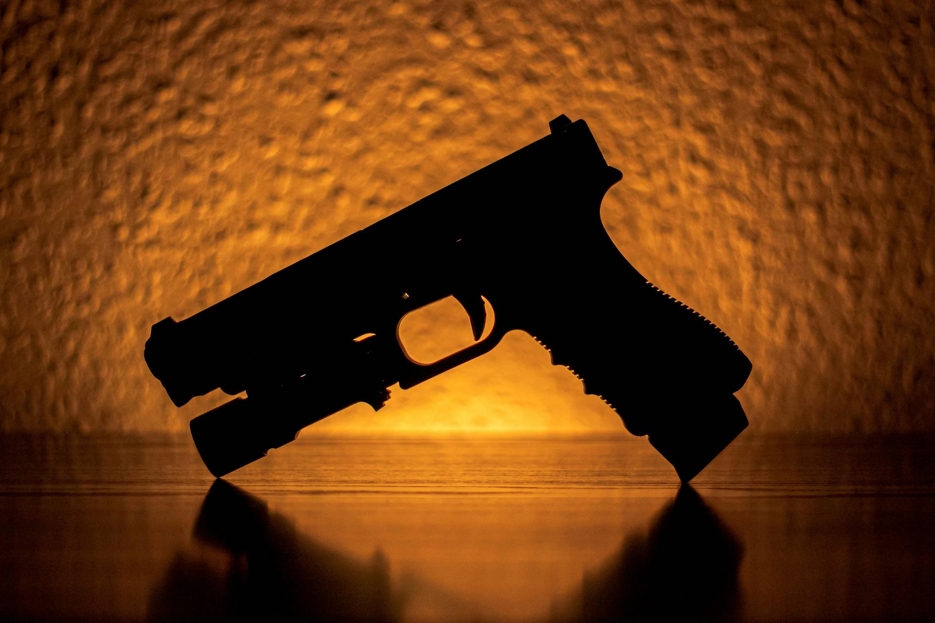 Silhouette of a handgun against a warm orange background with a reflective surface below