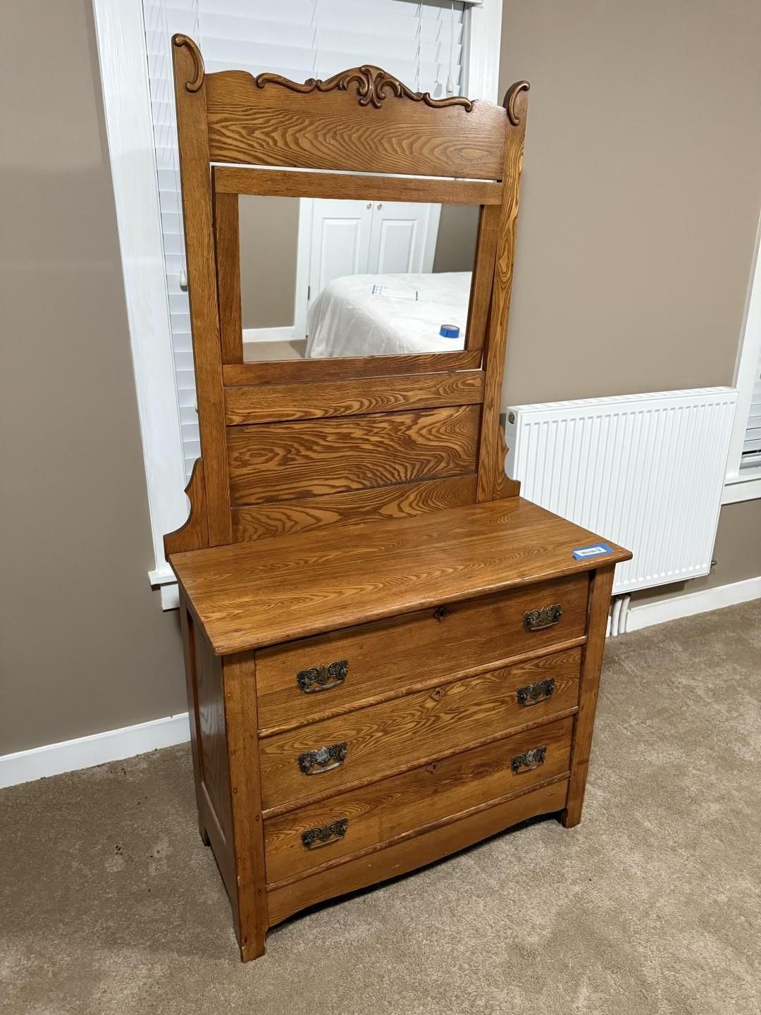 Wooden dresser with mirror, three drawers, ornate carving, in a room with a window and carpet.