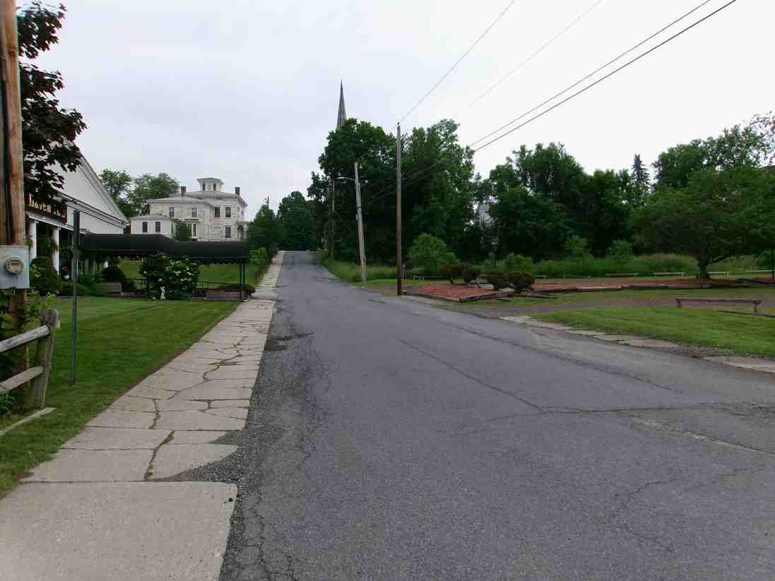 Empty suburban road beside a sidewalk and grassy lot, with houses and trees under an overcast sky