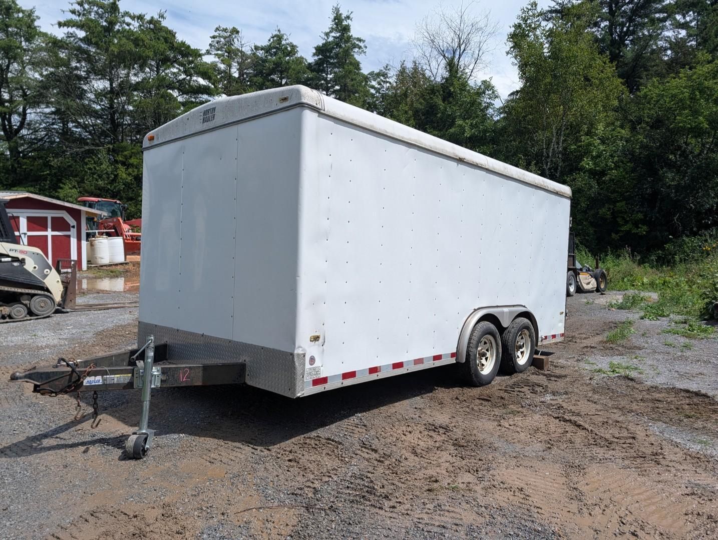 White enclosed cargo trailer parked on gravel with a tow hitch, outdoors near trees and sheds