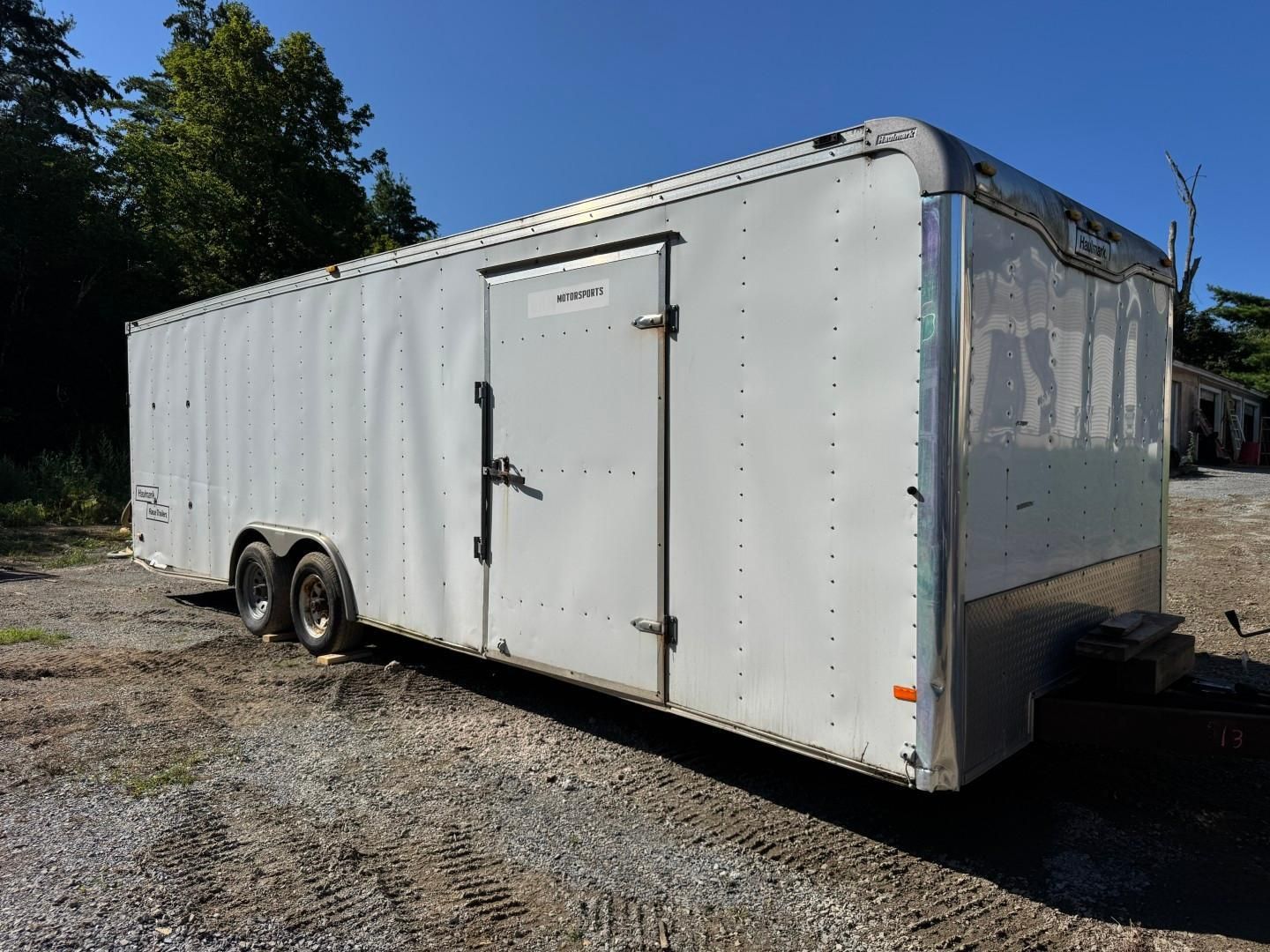White enclosed cargo trailer parked on gravel beside trees, with double rear wheels and side door.