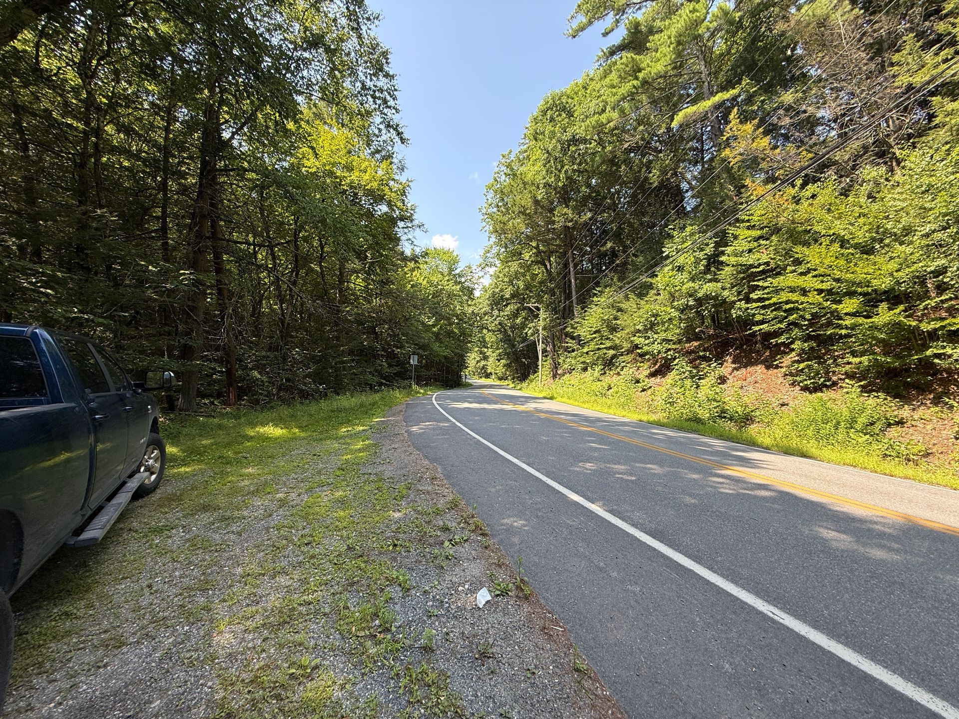 Narrow rural road curving through dense green trees, with a parked dark car on the left shoulder.