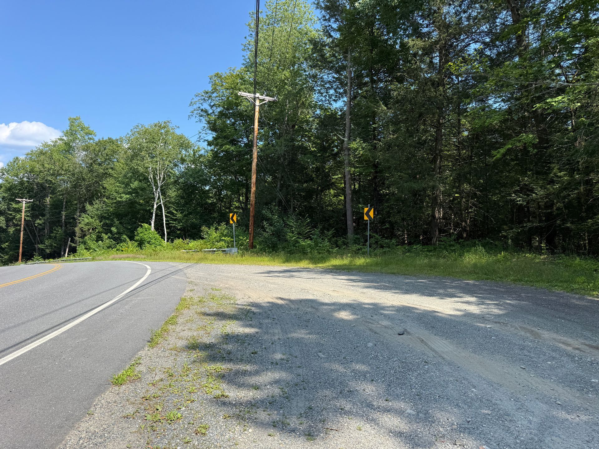 Curving road beside a gravel pull-off and dense green trees under a bright blue sky