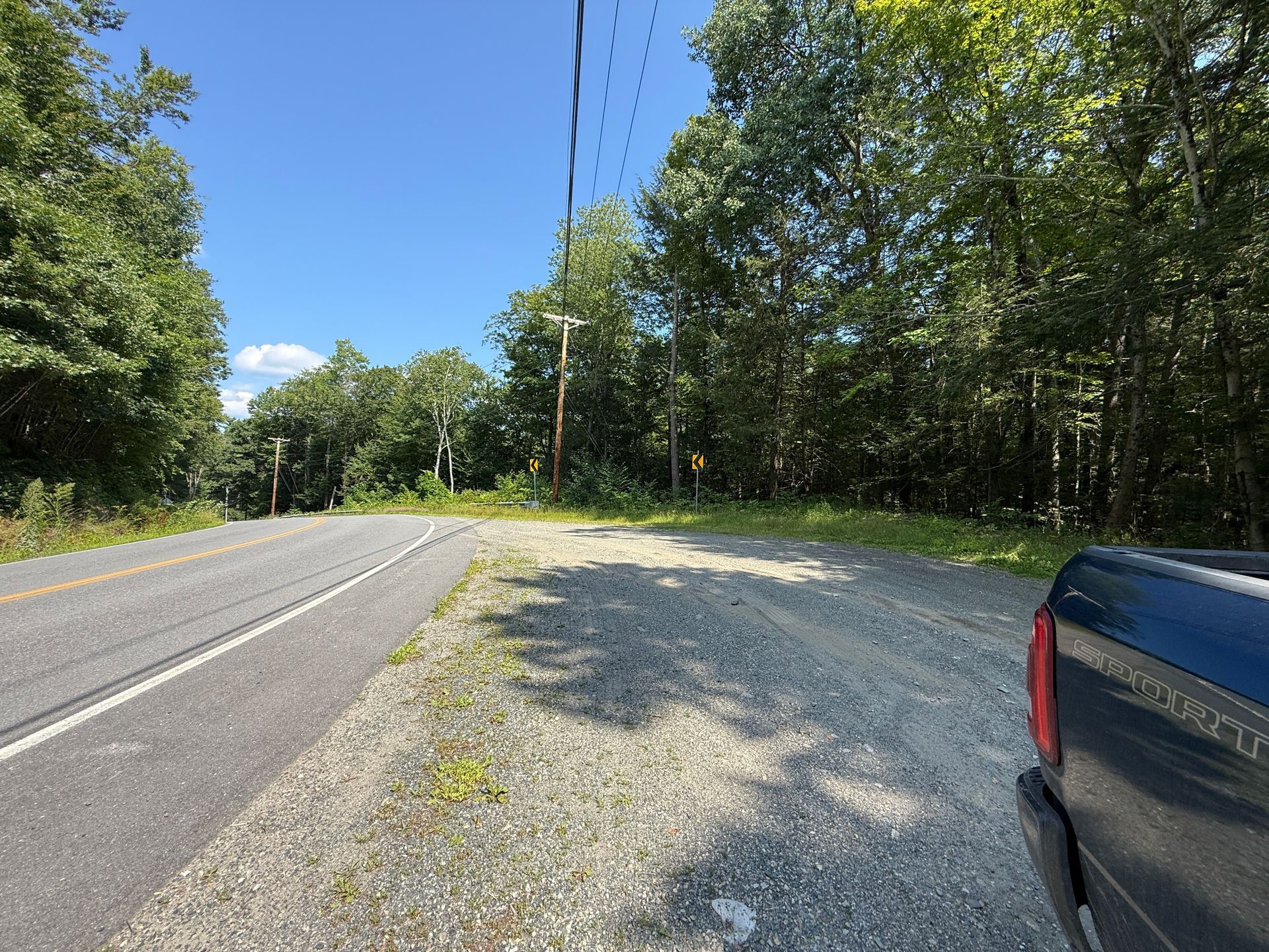 Rural roadside with trees, gravel turnout, and a parked dark pickup beside a two-lane road under clear sky