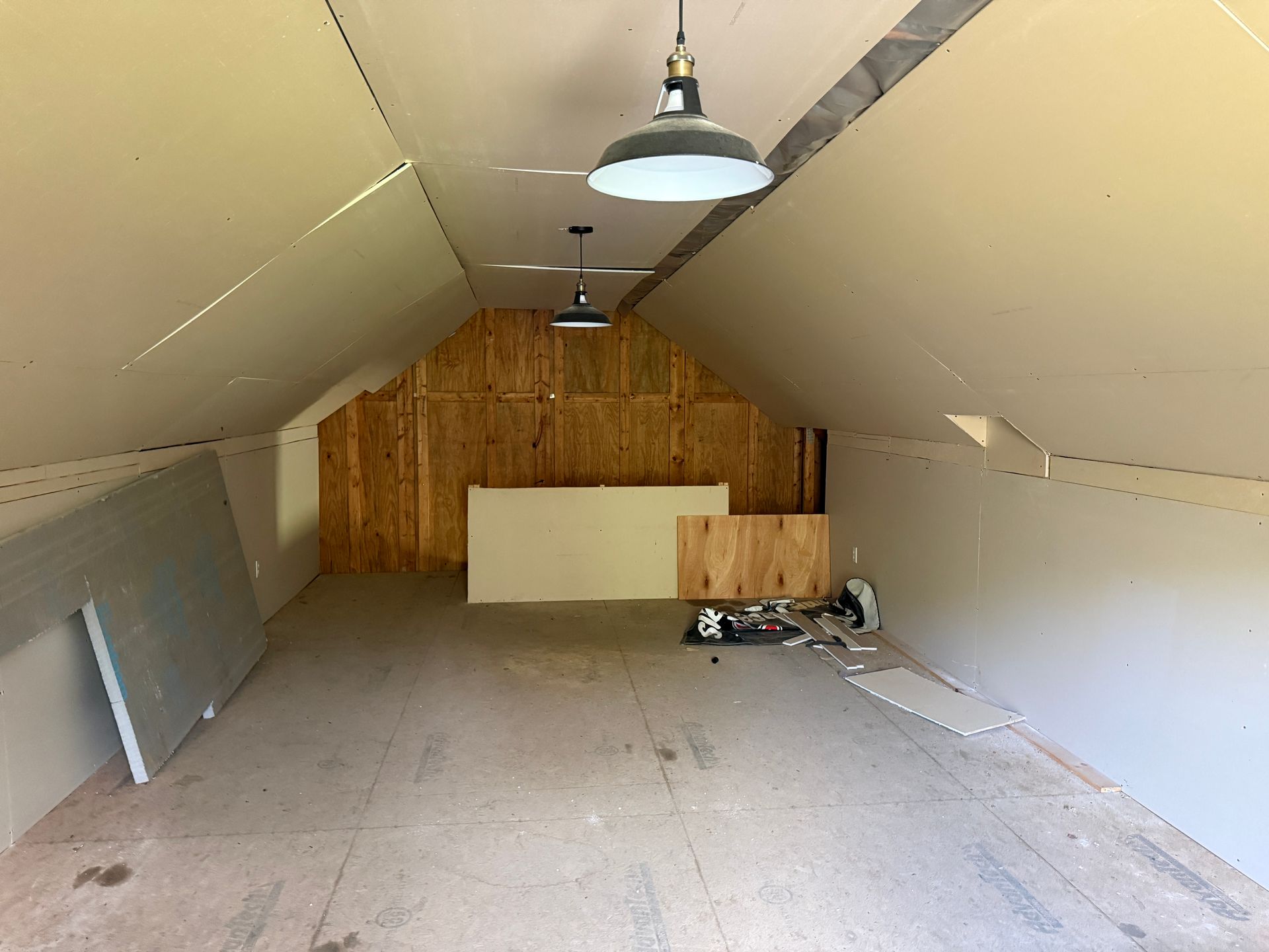 Empty attic room with sloped white walls, exposed wood back wall, and hanging lights.