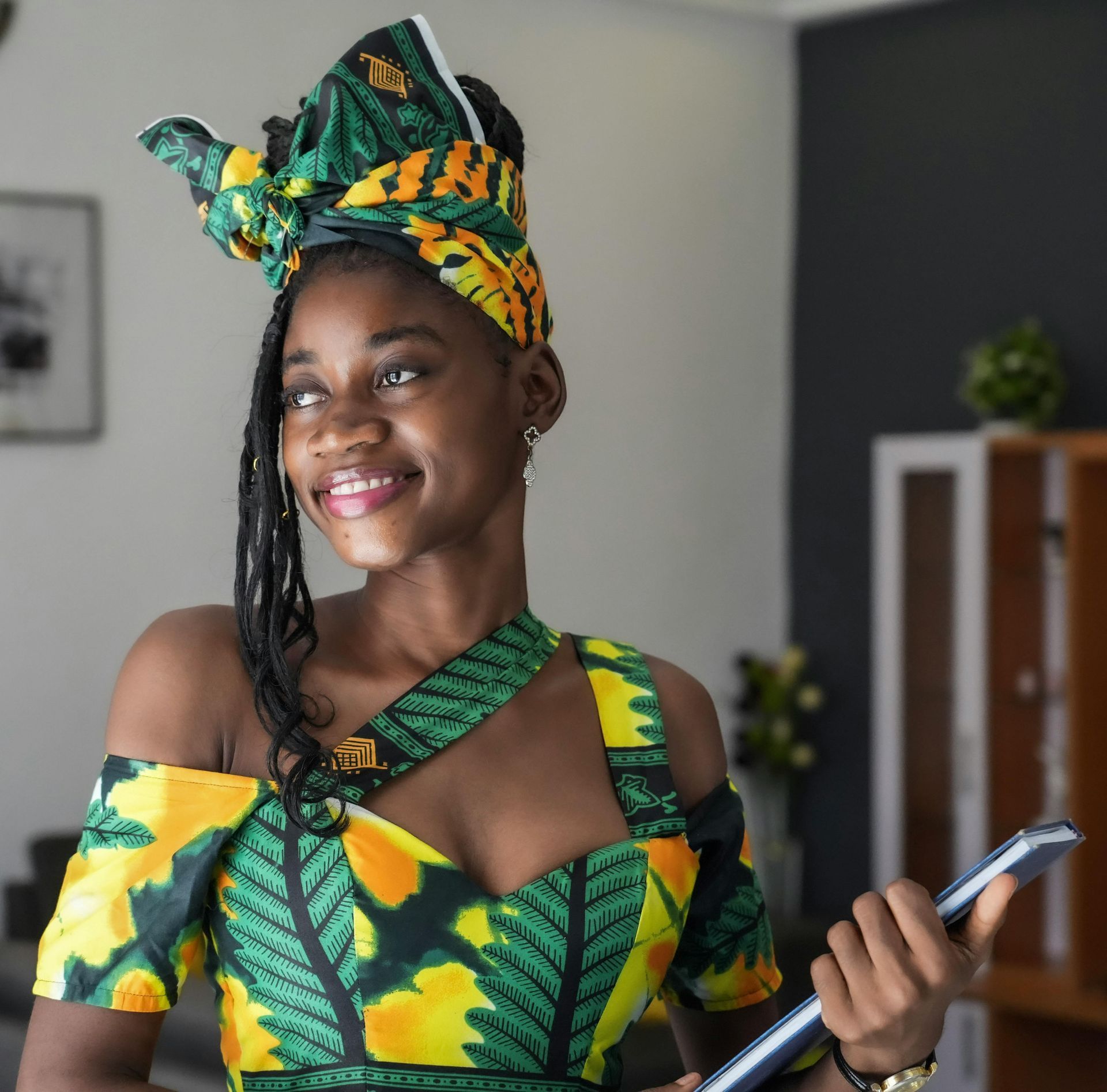 Femme en robe imprimée africaine verte et jaune et foulard souriant, tenant une tablette.