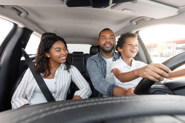Three people smiling in a car, with the driver steering and passengers wearing seatbelts.