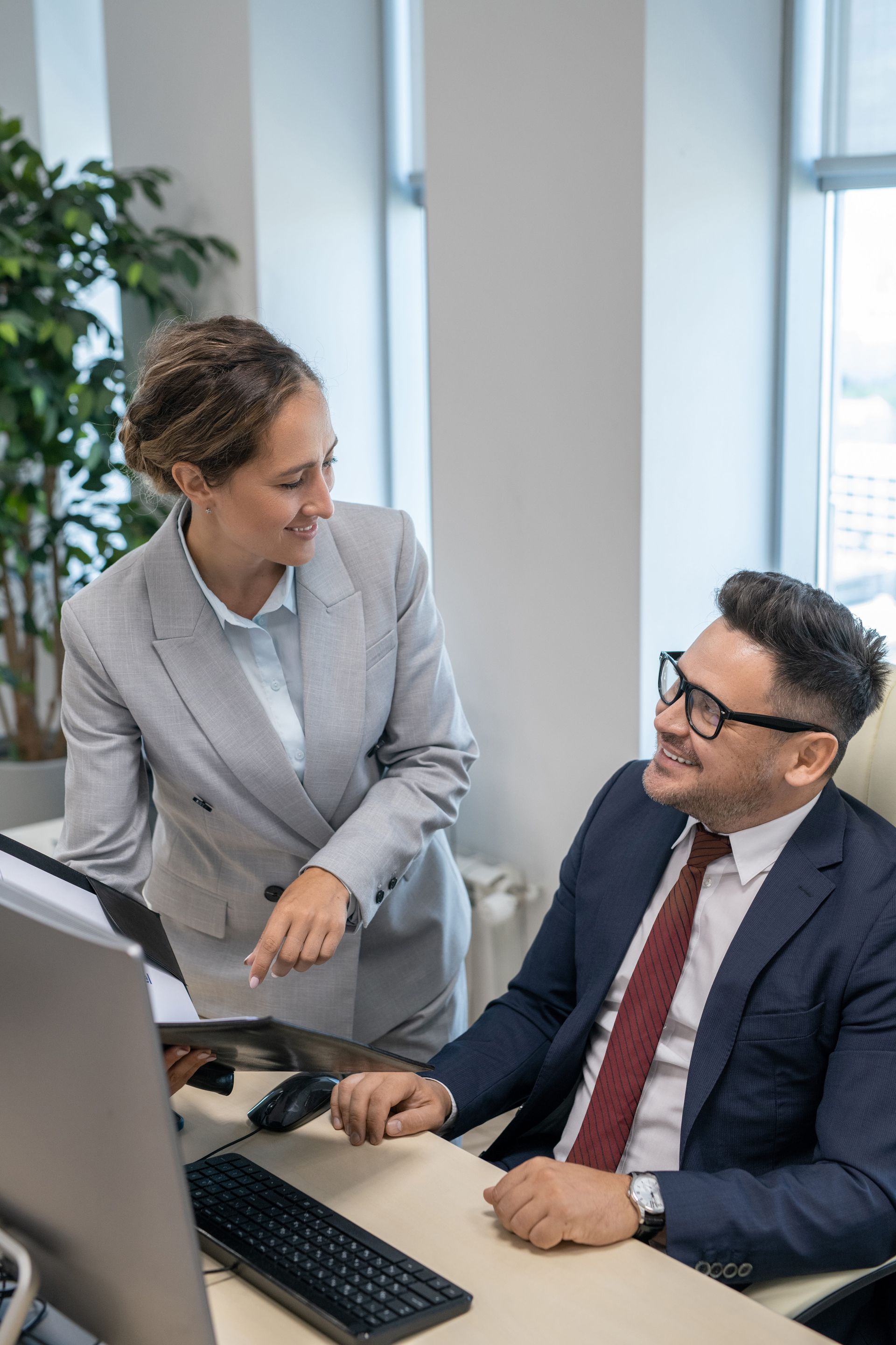 Two coworkers in business attire discussing at a desk in a bright office, smiling at a computer.