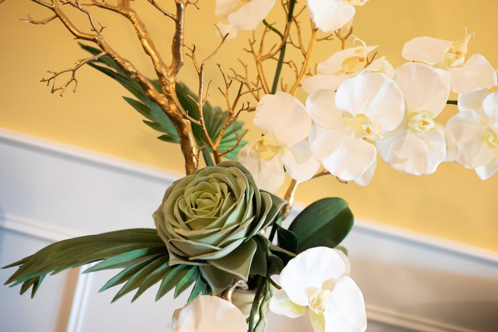 White orchids and a green rose in a floral arrangement with gold branches, against a cream wall.