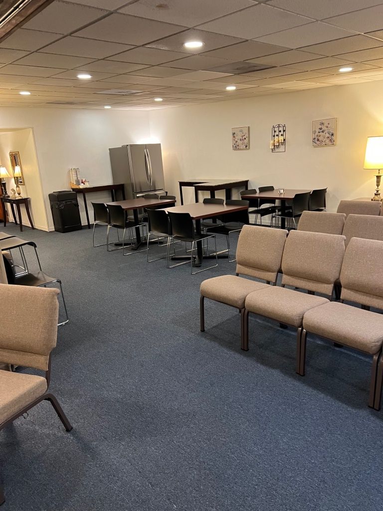 Meeting room with rows of chairs facing tables and chairs. Dark blue carpet, white walls.