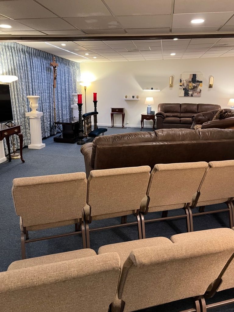 A funeral home interior with rows of chairs, a crucifix, and couches; blue carpet and cream walls.