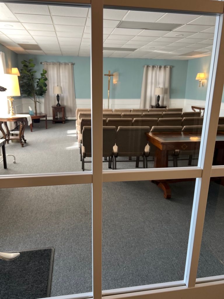 Interior of a funeral home chapel with rows of chairs, a podium, and a viewing area. Light blue walls and soft lighting.