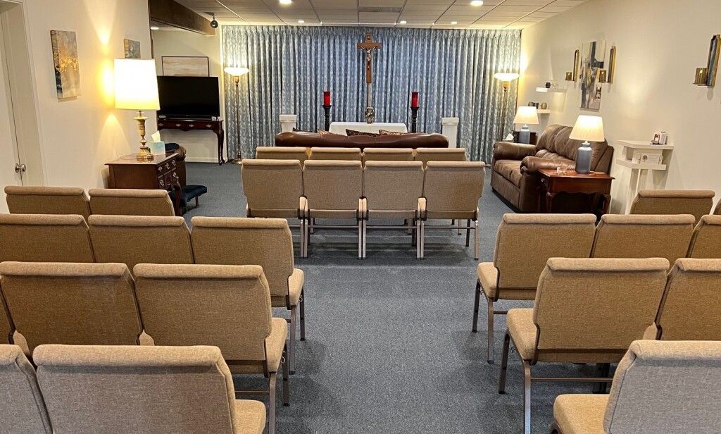 A funeral chapel with rows of tan chairs facing a raised altar with a crucifix, behind a silver curtain.