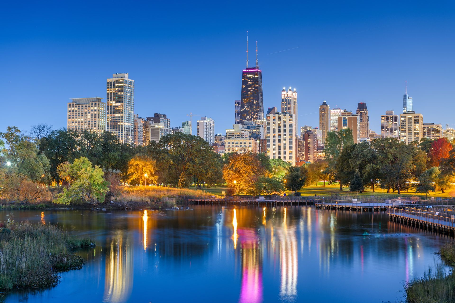 Chicago skyline reflecting in a lake at dusk, with trees in the foreground.