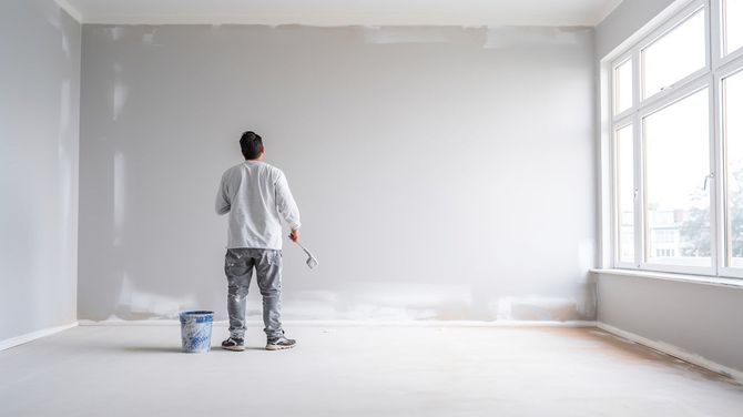 Person painting a white wall in a bright, empty room beside a large window.