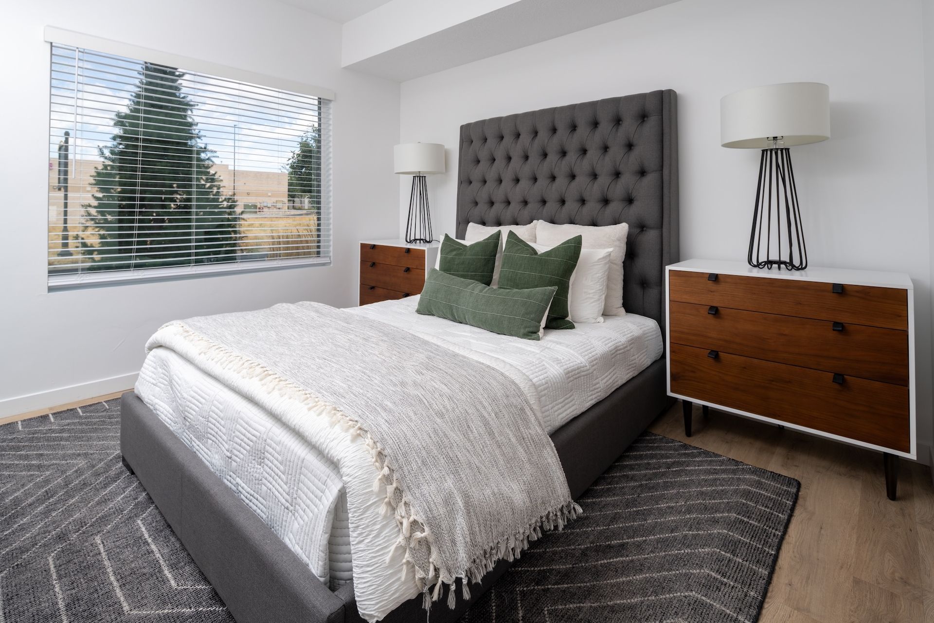 Bedroom with gray tufted headboard, two wooden nightstands, bed with white and green pillows, and a window.
