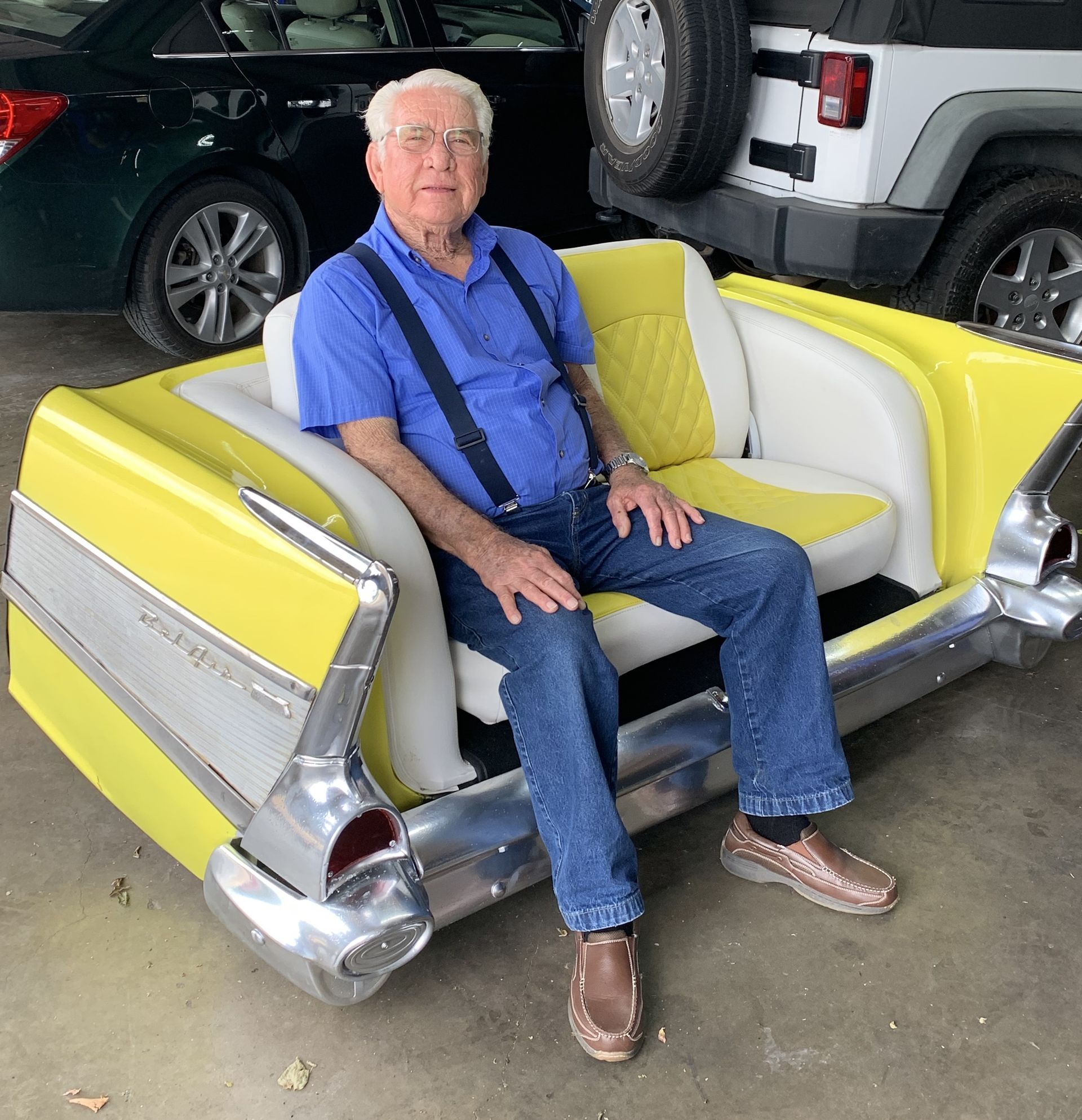 Man sitting on a bright yellow vintage car seat in a garage, wearing a blue shirt and jeans.