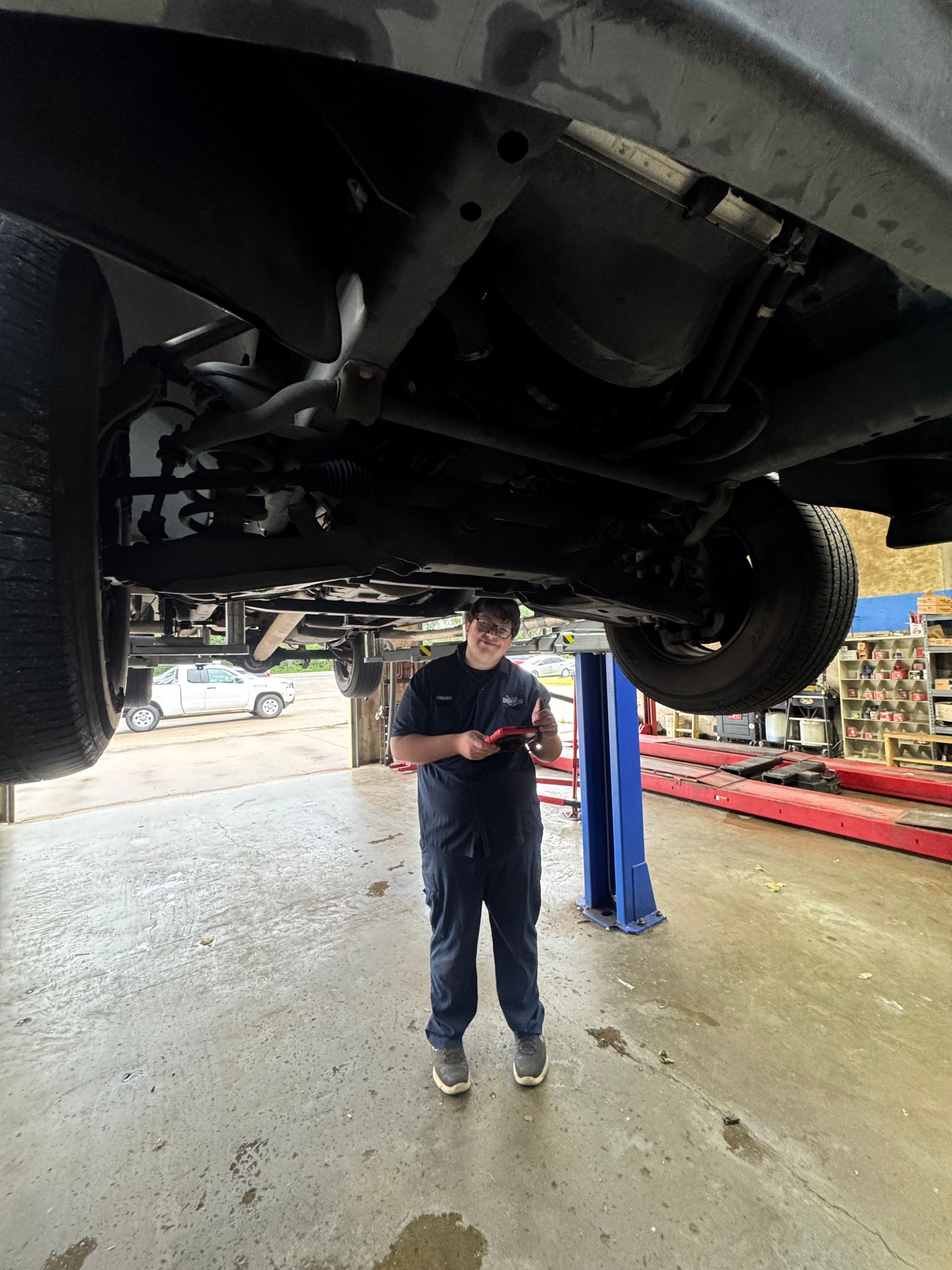 Mechanic standing under a raised vehicle in an auto shop, holding a phone beside a lift.