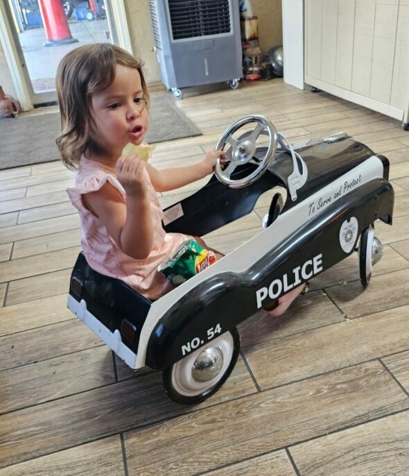 Child sitting in a toy police car, holding food, with a wooden floor and indoor background.