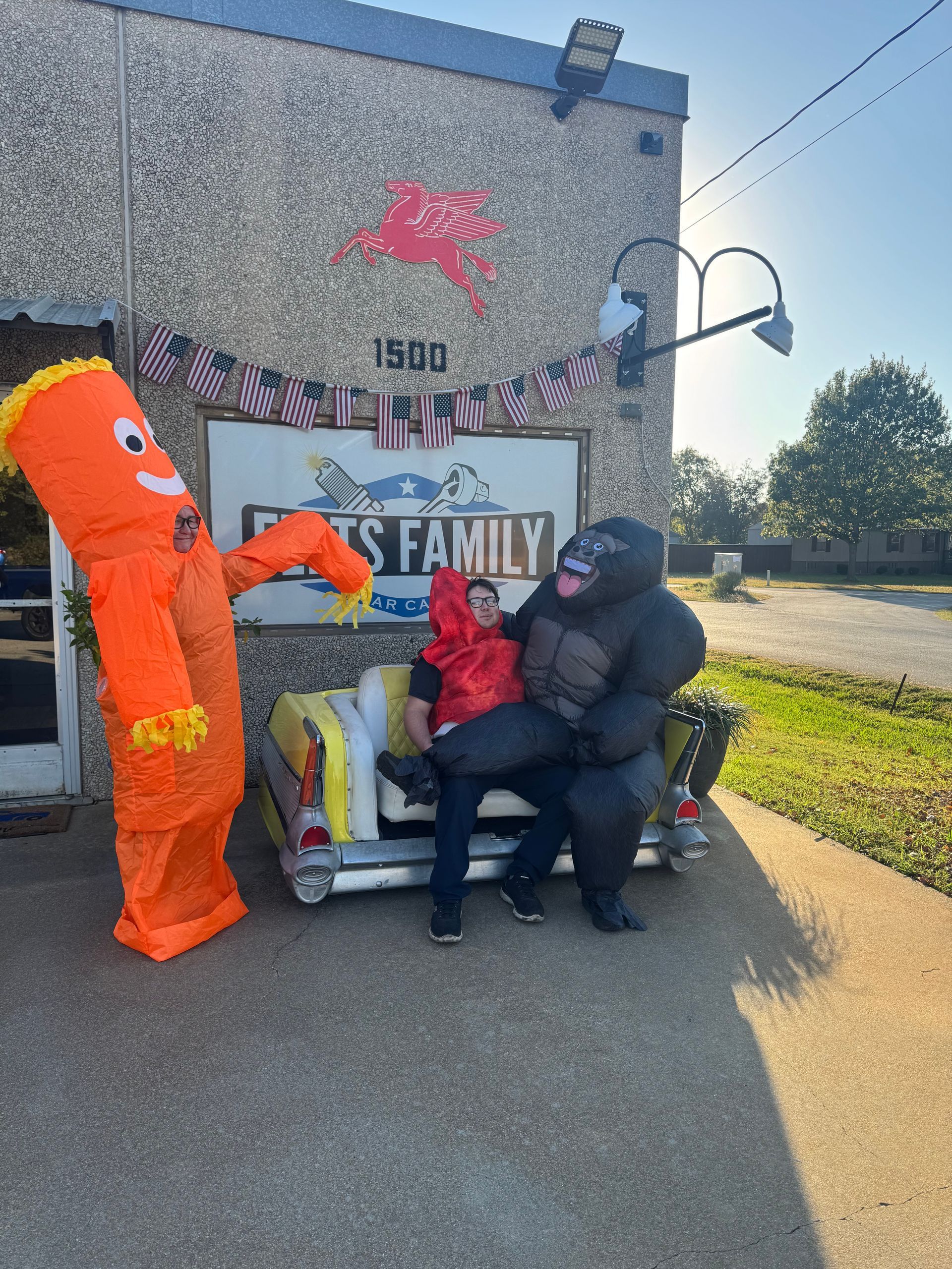 Two people seated on a couch with a large orange character standing beside them outdoors near a decorated building.