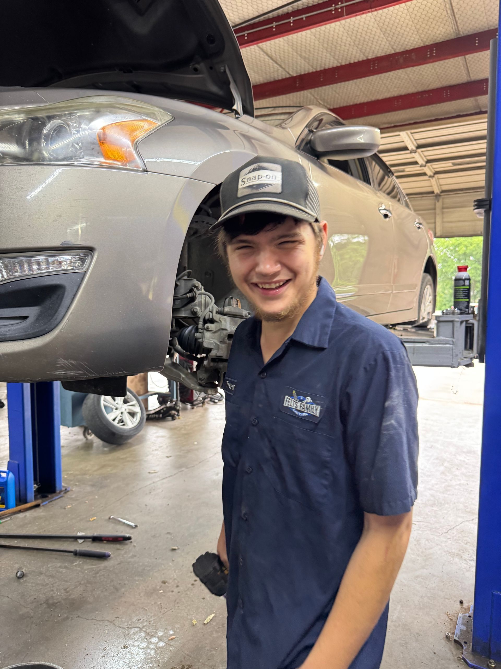 Mechanic smiling beside a car lifted in a garage, wearing a navy work shirt and cap