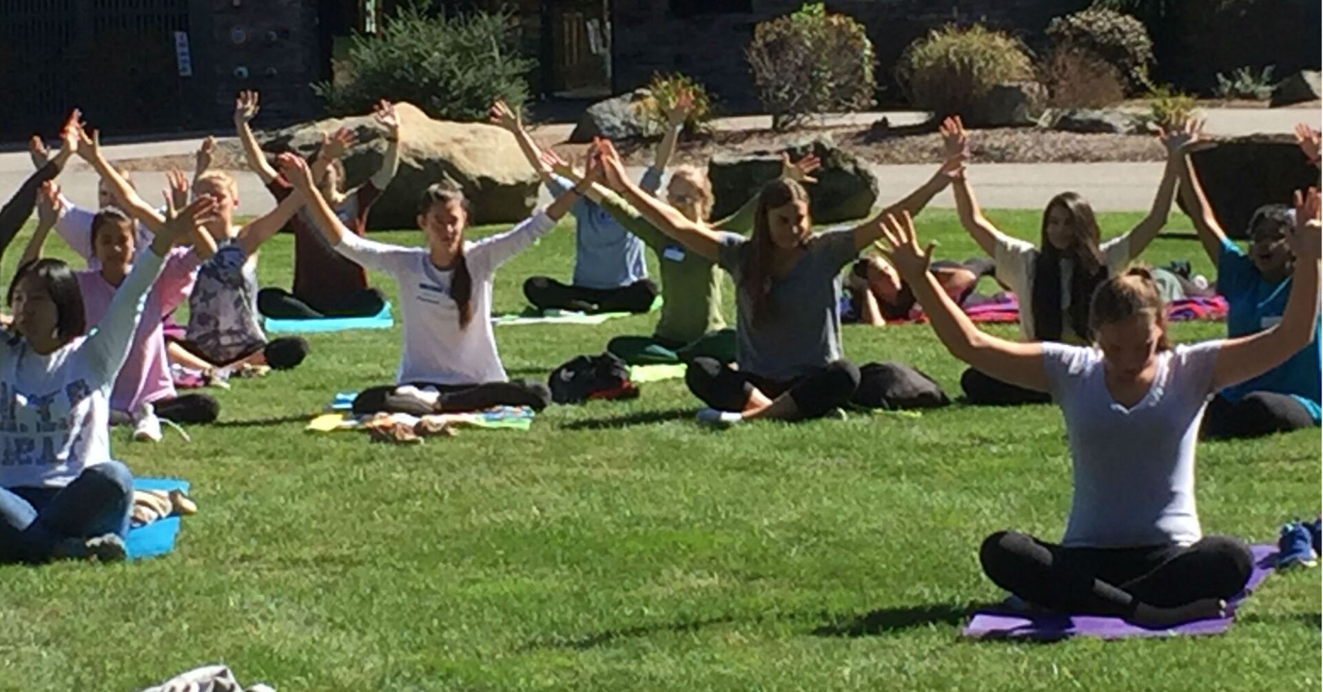 group of students outside with their arms up while sitting on mats on the grass 