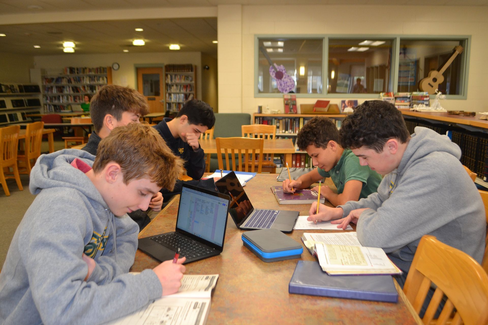 group of 5 students on laptops at a desk in the library with bookshelves in background