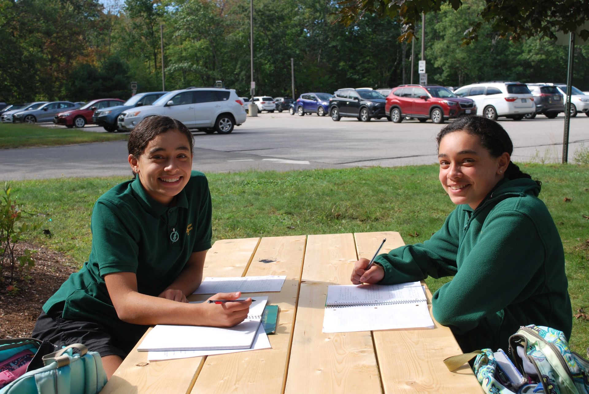 2 female students at a picnic table outside with grass and cars in the background