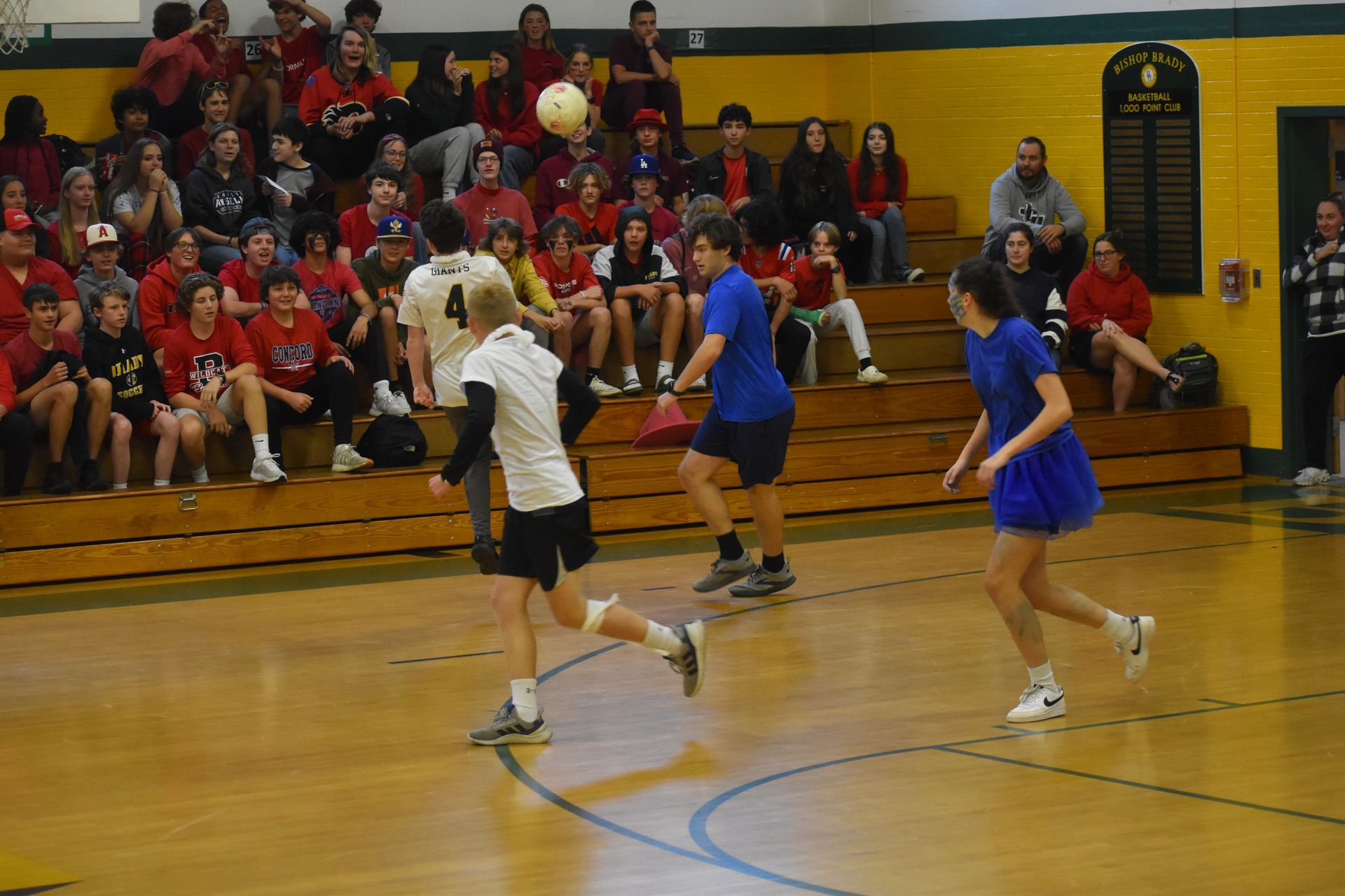 4 students playing basketball in the gym with many students wearing red in the background on bleachers