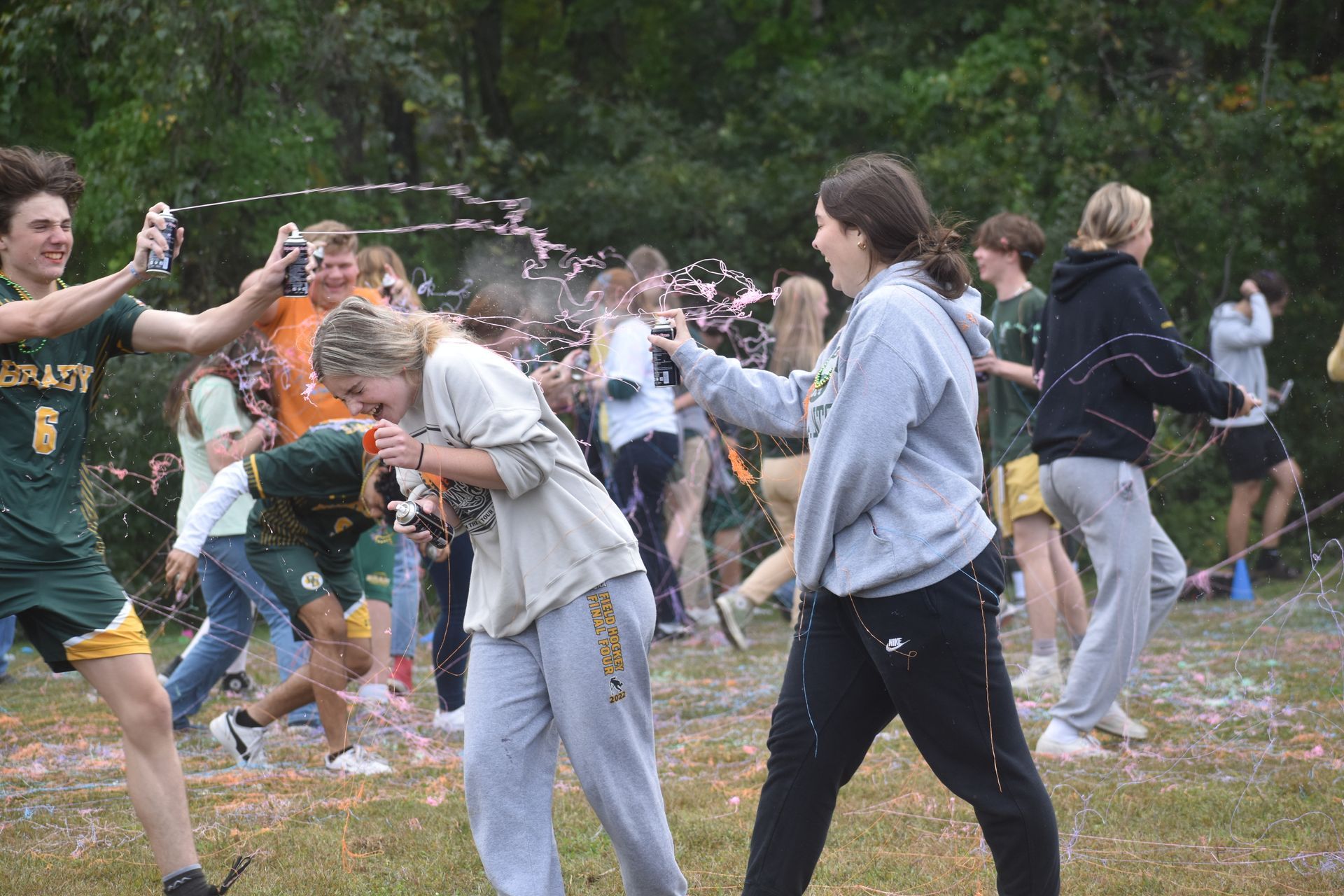 group of students spraying silly string outside on a field with trees in the background