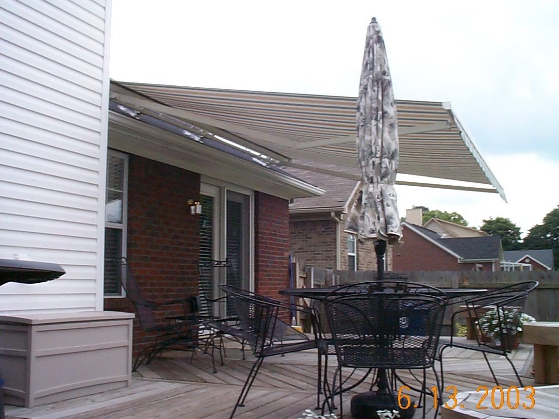 A brick patio with outdoor furniture and a closed umbrella under a large, striped retractable awning attached to a house.