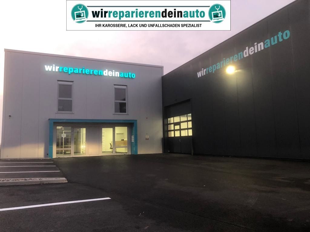 Außenansicht einer modernen Autowerkstatt in der Abenddämmerung mit beleuchtetem Eingang und Parkplatz.